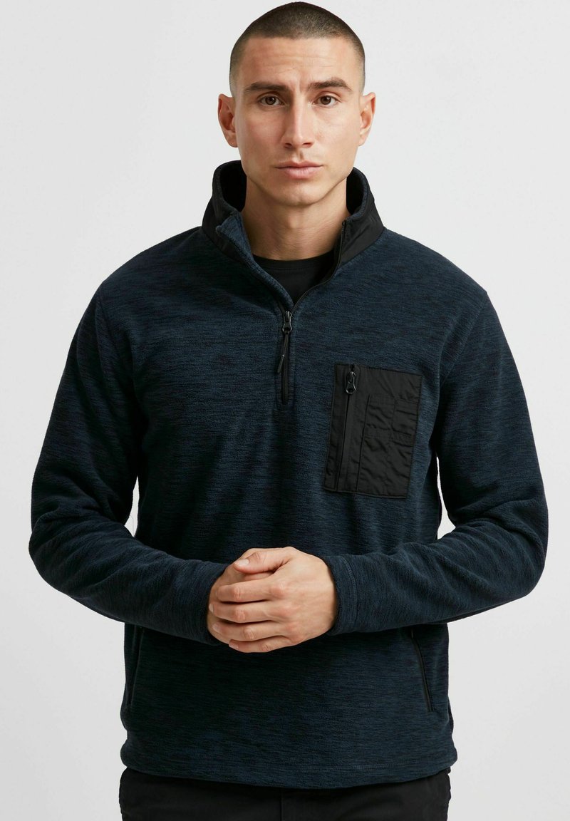 Dunkelblauer Fleece-Pullover mit einem Reißverschlusskragen, strukturiertem Stoff und einer schwarzen aufgesetzten Tasche auf der linken Brust. Lange Ärmel mit Bündchen.
