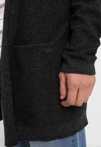 Hand ruht neben der dunkelgrauen, strukturierten Strickjackentasche mit einem kleinen schwarzen Markenetikett, getragen über blauen Jeans und einem weißen Hemd mit braunen Streifen.