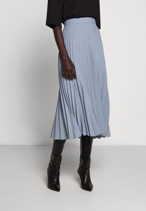 Person iført en lyseblå plisseret midi-nederdel, sort top og sorte knælange spidse støvler, stående mod en ensfarvet baggrund.