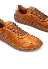 Sneaker in pelle marrone con punta perforata, cuciture a contrasto e lacci marroni. Presenta una suola intermedia texturizzata e accenti di logo discreti.
