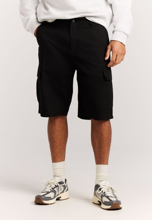 Persoon draagt zwarte cargoshorts, witte sokken tot aan de kuit, grijs-witte hardloopschoenen en een lichtgekleurde top met lange mouwen, staat tegen een effen achtergrond.