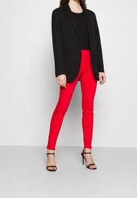 Pantalones ajustados rojos combinados con una americana negra y una blusa negra. El atuendo presenta un diseño minimalista en hardware y un corte elegante y ajustado.