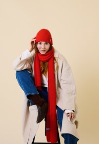 Red ribbed beanie and matching scarf paired with a long beige coat, blue jeans, and brown suede boots. Smooth, casual textile and layered design.