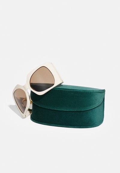 Gafas de sol blancas de gran tamaño con lentes tintados en marrón reposando sobre un estuche de terciopelo verde oscuro, con forma redondeada y textura suave.