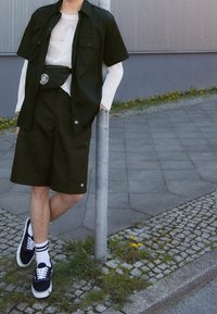 Chemise noire à manches courtes, haut en maille blanche ample, short foncé, sac banane noir et baskets noires avec semelles blanches et chaussettes à rayures.