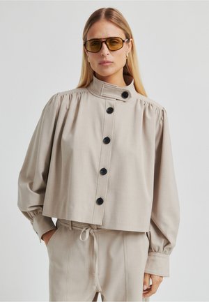 Chaqueta corta beige con cuello alto, botones negros y mangas fruncidas. Combinada con pantalones a juego, creando un conjunto coordinado.