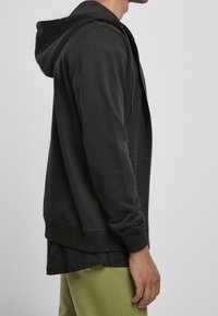 Sudadera con cremallera negra hecha de una mezcla de algodón, con un ajuste holgado, bolsillo tipo canguro y puños acanalados. Llevada sobre una camiseta negra y pantalones verdes.