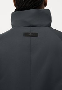 Άτομο που φαίνεται από πίσω, φοράει σκούρο γκρι μπουφάν με ψηλό γιακά και μικρό μαύρο λογότυπο Adidas στην επάνω πλάτη.