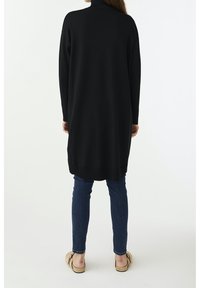 Tunic nera a maniche lunghe in maglia con colletto alto, caratterizzata da un fit comodo e orlo a coste, abbinata a jeans blu e scarpe beige.