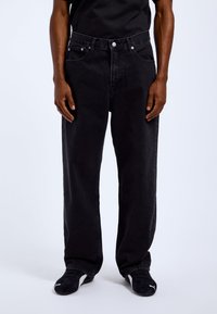 Black denim jeans with a relaxed fit, five-pocket design, button closure, and subtle stitching details. Worn with black athletic shoes.