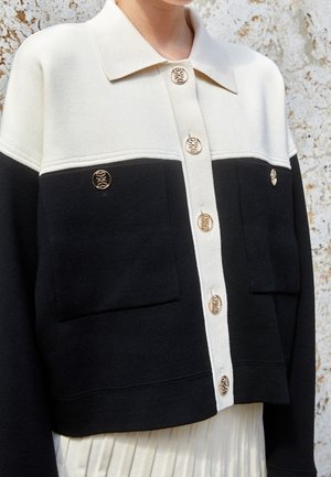 Dwukolorowy czarno-biały kardigan na guziki ze złotymi ozdobnymi guzikami i dużymi naszywanymi kieszeniami, noszony na plisowanej białej spódnicy.