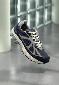 Αθλητικό παπούτσι από αεριζόμενο δίχτυ σε μπλε και γκρι χρώμα με λευκά κορδόνια, λευκή και μαύρη σόλα, τοποθετημένο πάνω σε γκρι πάτωμα κοντά σε γυάλινους κυβάς.