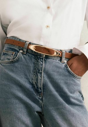 Rudas odinis diržas su lygia apdaila ir ovalia aukso sagtimi, dėvimas ant šviesiai mėlynų džinsų. Iš dalies matoma marškiniai su mygtukais.