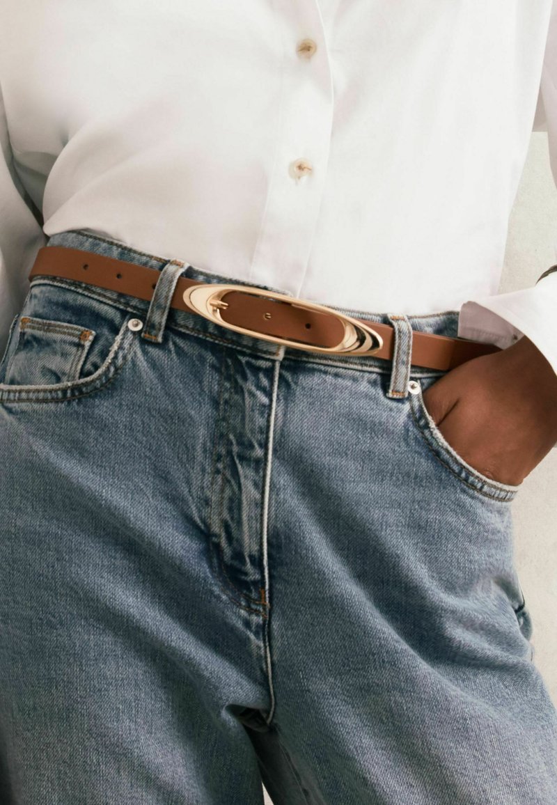 Brūns ādas jostas ar gludu apdari un ovālu zelta sprādzi, kas uzvilkta virs gaiši zila džinsu biksēm. Pogājama krekla daļa daļēji redzama.