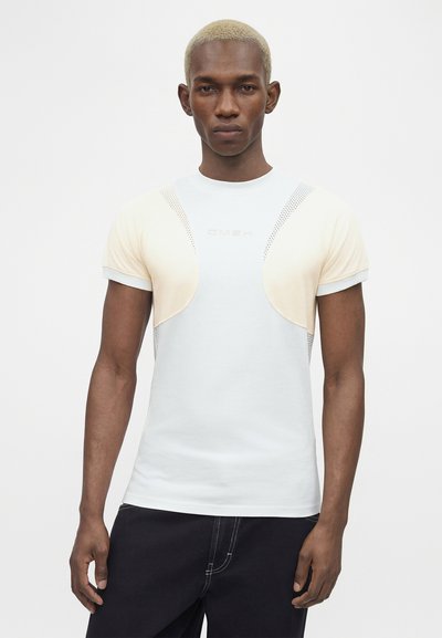 Άνδρας με κοντά ξανθά μαλλιά που φοράει λευκό μπλουζάκι με μπεζ δίχτυα, στέκεται μπροστά σε απλό φωτεινό φόντο.