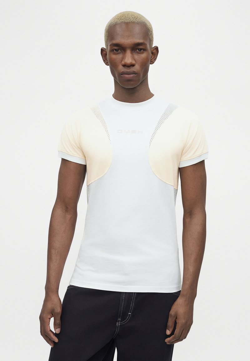 Homme aux cheveux blonds courts portant un t-shirt blanc avec des panneaux en maille beige, debout devant un fond clair uni.