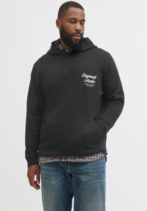 Man draagt een zwarte hoodie met witte tekst op de borst, daaroverheen een geruit overhemd en een blauwe jeans, staand tegen een effen achtergrond.