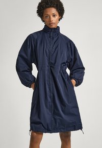 Manteau long navy en tissu léger, avec un col montant, des manches bouffantes et des poignets élastiques. Ourlet à cordon de serrage et poches latérales.