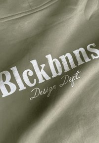 Olijfgroen stof met witte tekst: "Blckbnns" in vet lettertype, daaronder "Design Dept." in een cursieve stijl, met een gladde textuur.