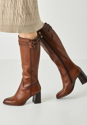 Brown leather knee-high boots with block heels, side zipper, and double buckle straps, worn with a beige knitted skirt.