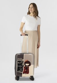 Valigia rigida con finitura lucida, caratterizzata da un design grafico di una ragazza con un cappello su uno sfondo diviso tra colori beige e marrone.