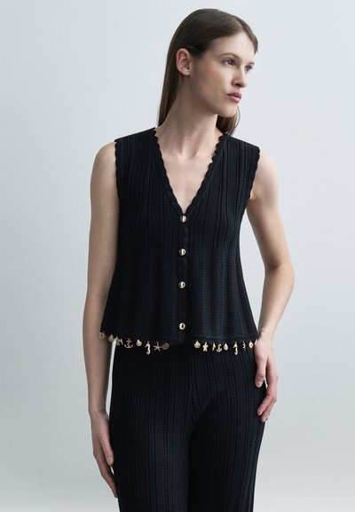 Femme portant un gilet tricoté sans manches noir avec des boutons et des charms dorés décoratifs sur le thème de la mer le long de l'ourlet, accompagné d'un pantalon assorti.