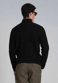 Pull en tricot noir à côtes avec un col montant, des poignets ajustés et un ourlet inférieur, associé à un pantalon vert olive. Le pull présente un motif texturé.