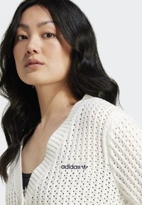 Cardigã branco tricotado com decote em V, apresentando um padrão texturizado e um logótipo bordado da Adidas em azul marinho no peito.