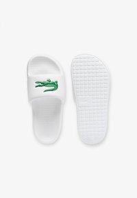 Witte slip-on sandalen met een groene krokodillenlogo op de band. Het materiaal is glad, met een gestructureerde zool die een gridpatroon heeft.
