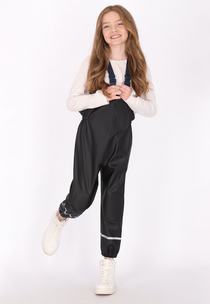 Šypsena mergaitė su ilgais plaukais, dėvinti juodas vandeniui atsparias kelnes ir baltais sportbačiais, pakėlusi vieną koją šiek tiek virš žemės, priešais baltą foną.