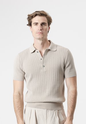 Hombre con cabello corto y ondulado, que lleva una camiseta polo de punto beige de manga corta y pantalones plisados de color claro, de pie frente a un fondo liso.