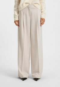 Helle beige weit geschnittene Hose aus glattem Stoff, mit vorderen Falten und klassischem Bund. Schwarze spitze Absatzschuhe runden den Look ab.