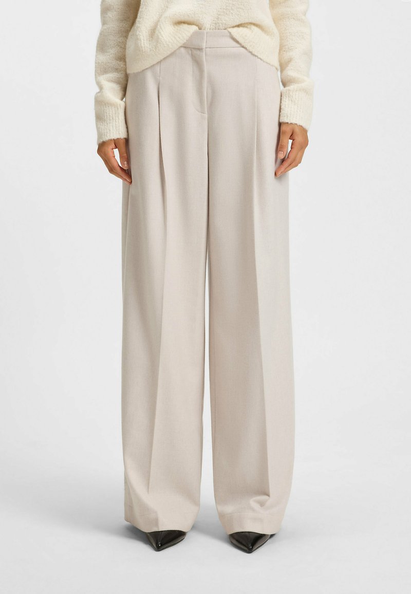 Helle beige weit geschnittene Hose aus glattem Stoff, mit vorderen Falten und klassischem Bund. Schwarze spitze Absatzschuhe runden den Look ab.