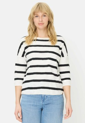 Kvinde med blondt hår iført sort-hvid stribet langærmet skjorte og lyseblå jeans, stående foran en ensfarvet hvid baggrund.