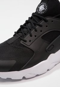 Black athletic sneaker with mesh and synthetic materials, featuring a textured toe box, prominent lacing system, and white sole.