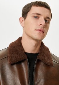 Giacca in pelle marrone con colletto soffice marrone, chiusura con zip e una texture liscia. Il design presenta un taglio semplice e classico.