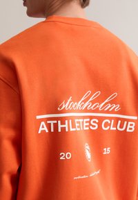 Pomarańczowa bluza z białym napisem: "stockholm ATHLETES CLUB 20 15" oraz logo. Materiał wydaje się miękki, z okrągłym dekoltem.
