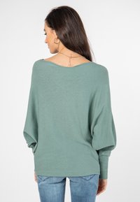 Losse, lange mouwen trui van zacht groene stof; voorzien van een brede halslijn en ribgebreide manchetten. Achterkant heeft een kettingaccent.