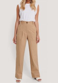 Beżowe spodnie o wysokim stanie z zakładkami z przodu, wykonane z mieszanki bawełny. Posiadają pojedyncze zapięcie na guzik oraz proste, szerokie nogawki.