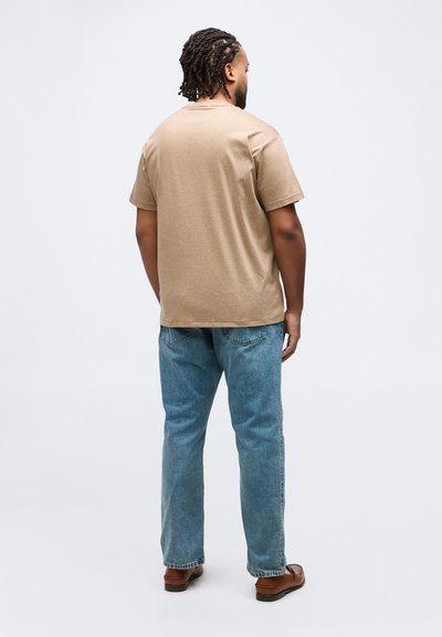 Άνδρας με πλεξούδες, γυρισμένος πλάτη, φοράει μπεζ μπλούζα, μπλε τζιν και καφέ δερμάτινα παπούτσια σε λευκό απλό φόντο.