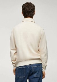 Suéter crema con cuello tipo polo, mangas largas y puños acanalados, combinado con jeans de denim azul con bolsillos traseros.