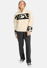 Pullover in pile crema con motivo a spirale nera, chiusura a mezzo zip e polsini neri a contrasto. Abbinato a pantaloni neri larghi e sneakers.