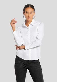 Weißes Hemd mit Knopfleiste aus glattem Stoff, mit spitzem Kragen, langen Ärmeln mit Manschetten und abgerundetem Saum. Kombiniert mit dunkler Jeans.