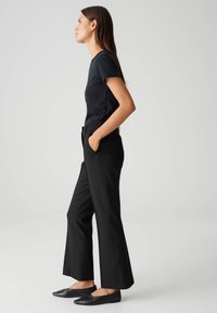 Zwart aansluitend T-shirt, high-waisted zwarte broek met een uitlopende pijp, en zwarte ballerina's, gedragen door een model in profiel tegen een effen achtergrond.