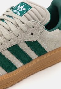 Gröna och grå Adidas Samba-sneakers med överdel av mocka, tre gröna ränder, en vadderad tunga och en sula av gummigummi med ett texturerat mönster.