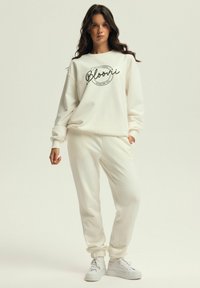 Helles Sweatshirt und Jogginghose aus weichem Stoff, mit einem runden schwarzen Grafikdruck auf der Vorderseite. Weiße Sneaker runden das Outfit ab.