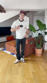 Bílá trička s černým logem, černé kalhoty a stříbrné boty. Vnitřní prostředí s hnědou sedačkou, rostlinou a dřevěnou podlahou.