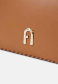 Borsa in pelle marrone con un logo metallico dorato a forma di "A" con un piccolo elemento decorativo. Texture liscia e design elegante.