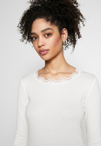 Vit långärmad topp med ribbad struktur och en scallop-lace dekoration längs halsringningen. Åtsittande design.