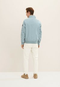 Giacca bomber azzurro chiaro con colletto alto e polsini a costine, indossata su pantaloni bianchi. La giacca presenta un piccolo logo del marchio sulla manica.